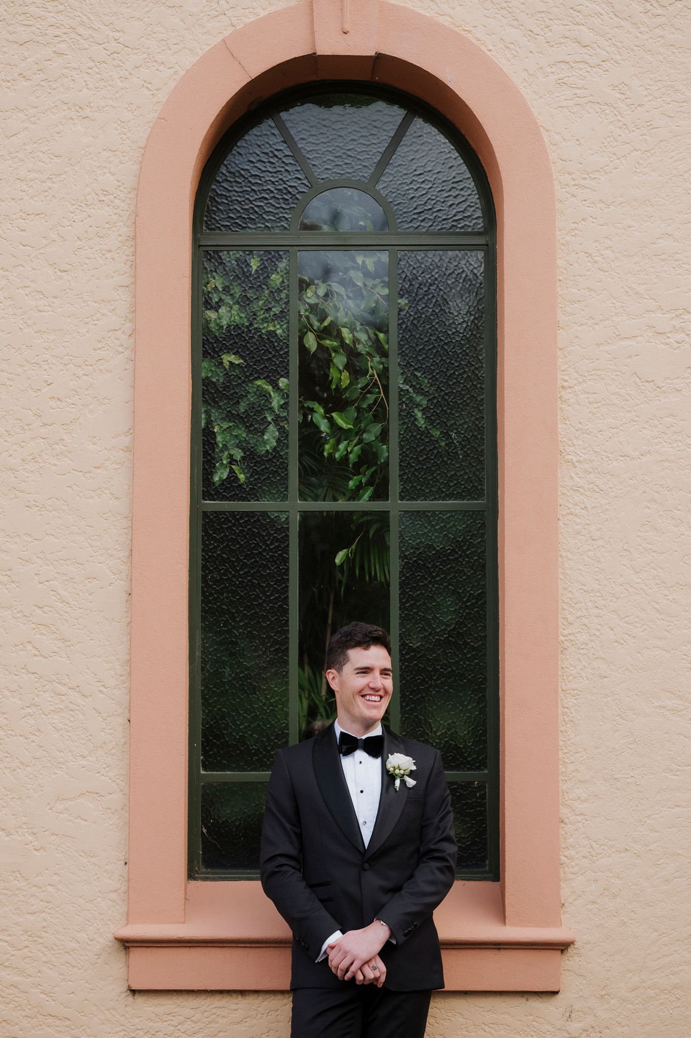 A groom in a black tuxedo stands alone in front of a pink arched window.