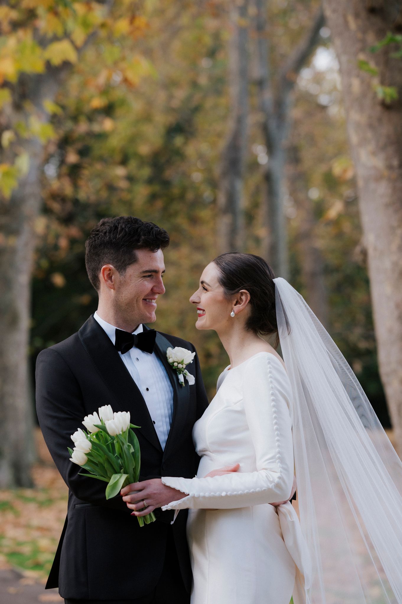 A couple shares a joyful moment among autumn trees while holding white flowers.