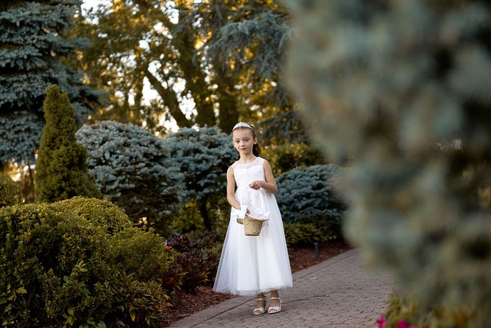Child in white dress carrying basket walking along garden path at sunset.
