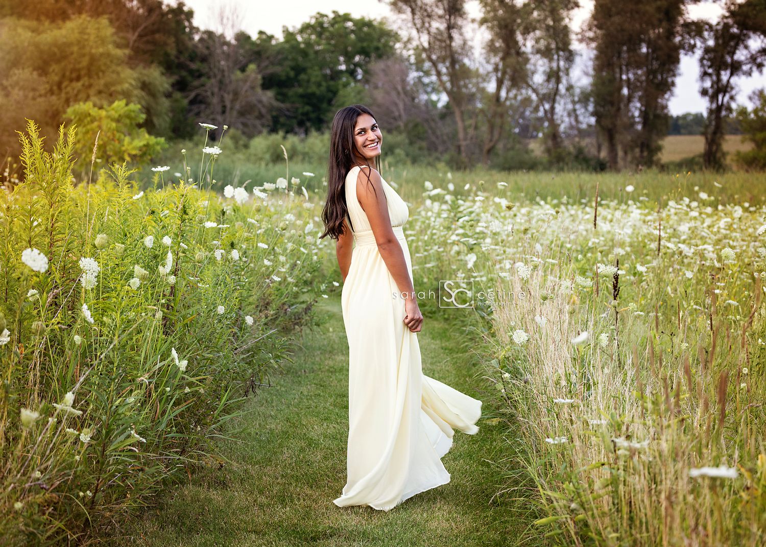 Someone in an elegant white wedding dress stands in a wildflower meadow with tall grass and daisies at sunset.