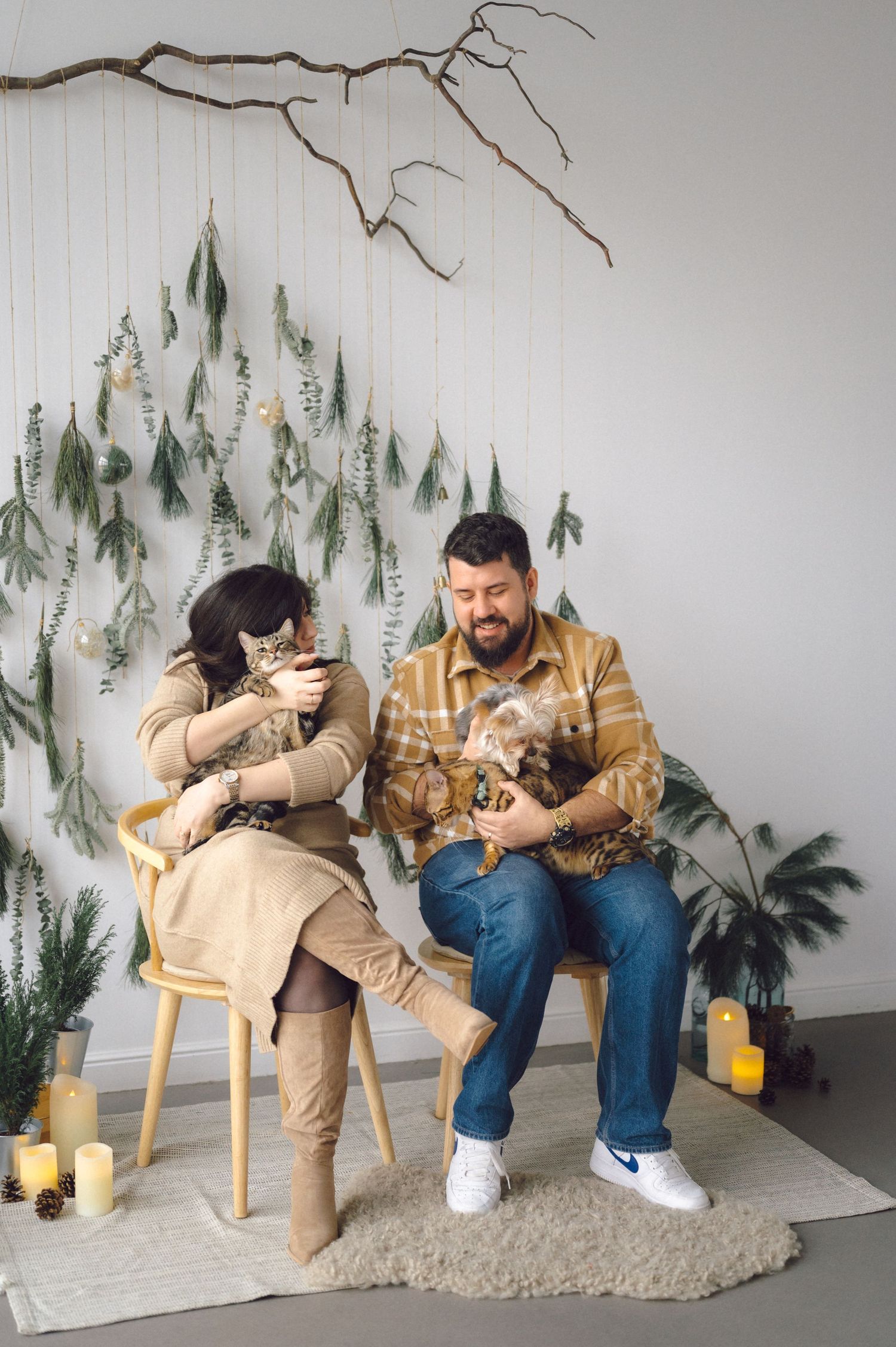 Two people sit on wooden chairs in a cozy corner decorated with hanging evergreen branches and holding pets.