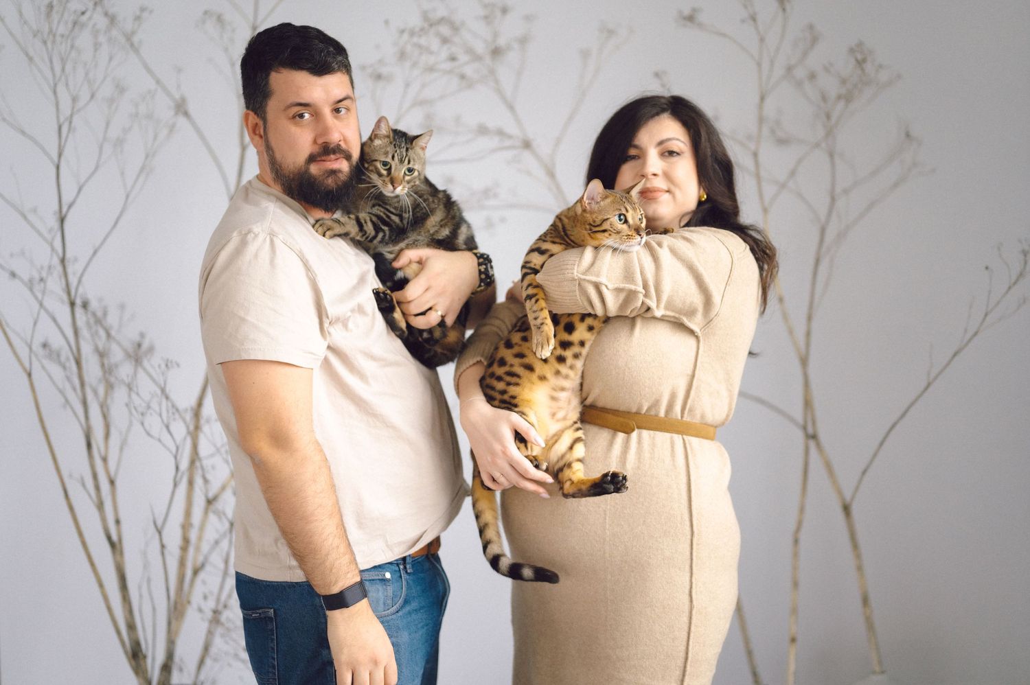 A couple cradles two tabby cats while posing in a maternity photo session against a pale background.
