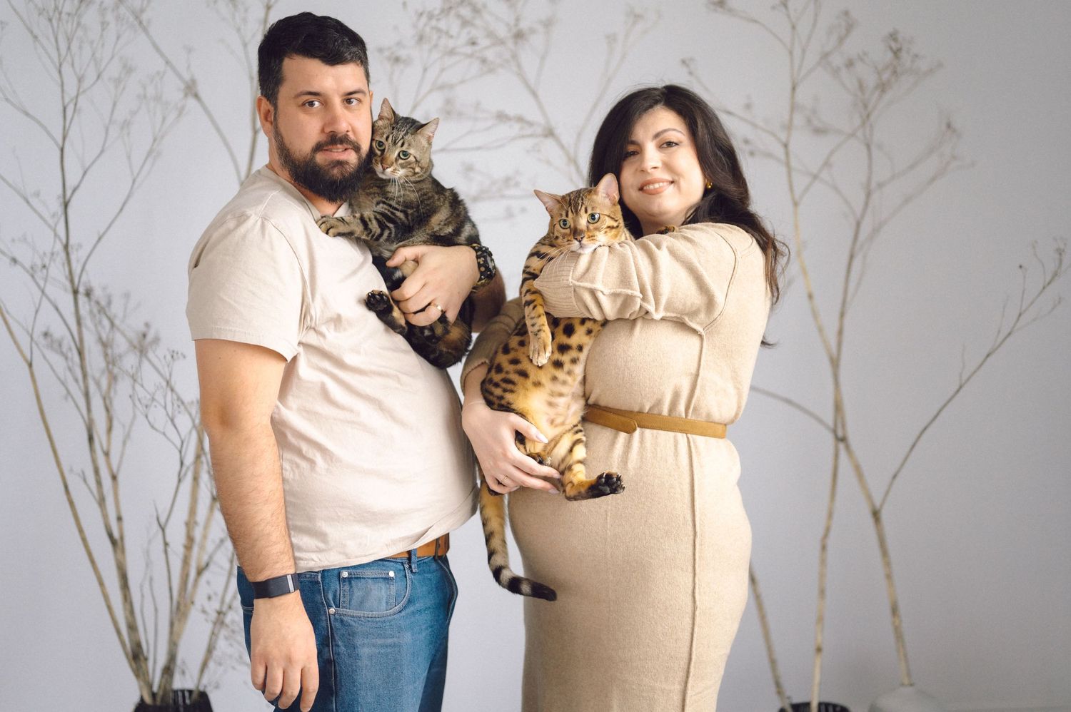 Maternity photo series showing couple holding tabby cat while posing against light gray backdrop.