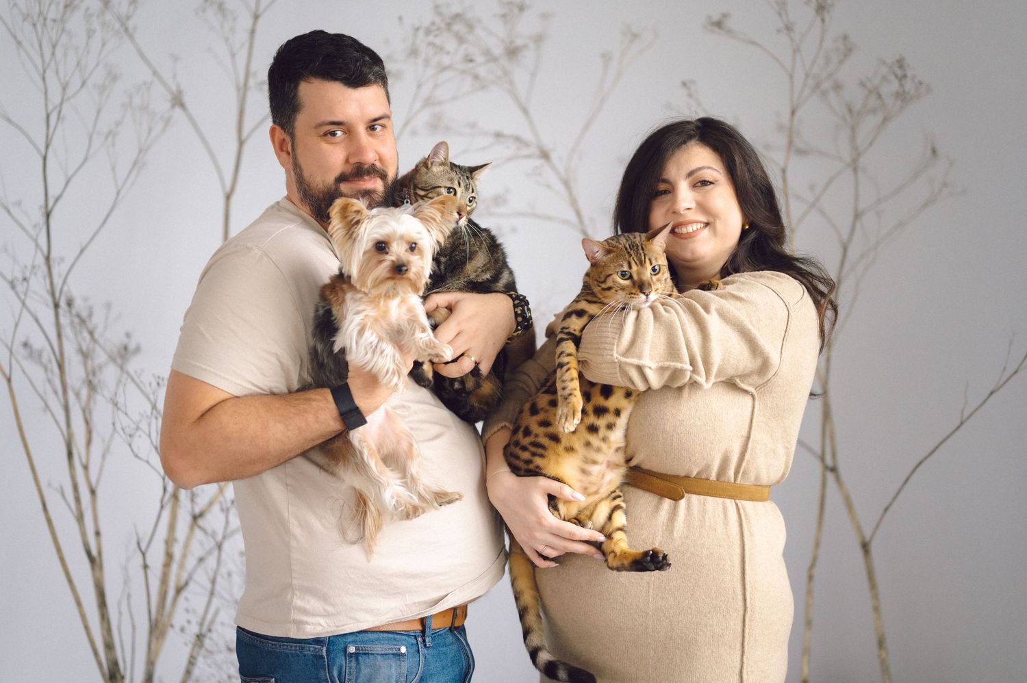 A couple holding a small dog and giraffe plush toys pose together in a cozy lifestyle photoshoot with white branches.