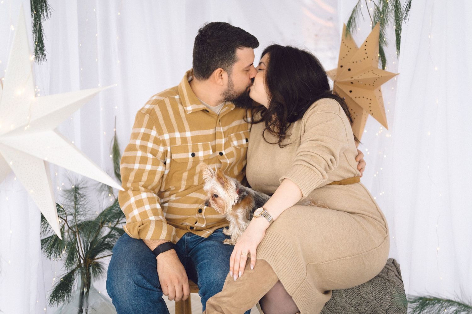 Young couple wearing neutral colored clothing sit together and embrace near plants and white wall during photo session.
