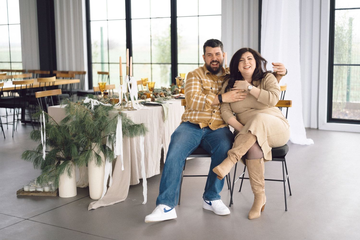 Two people pose casually on chairs in a bright modern event venue with elegant table settings and greenery.
