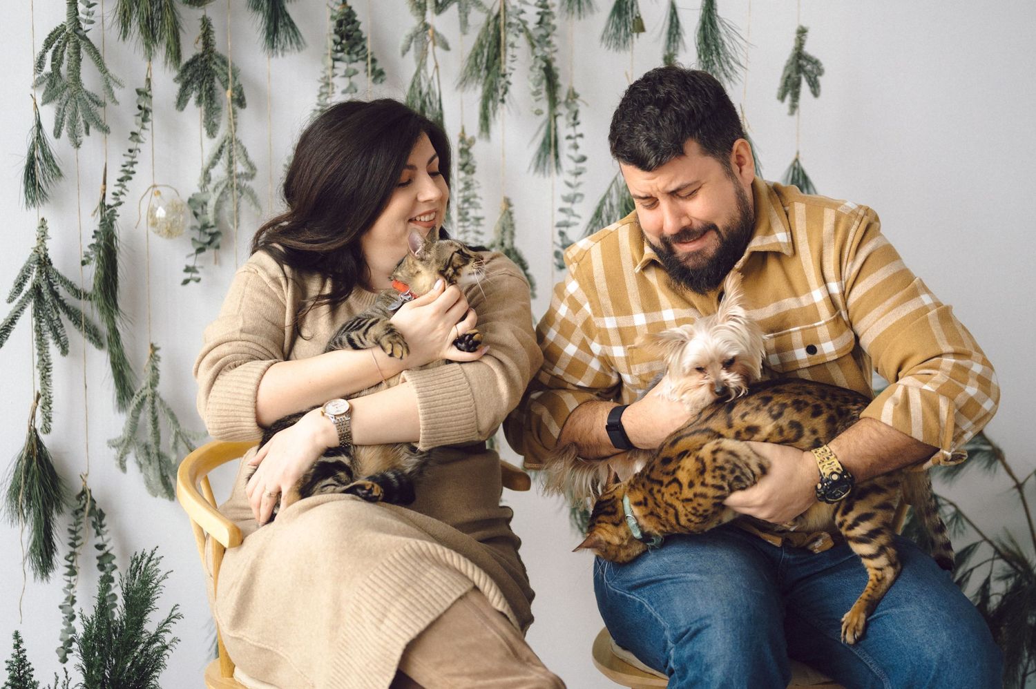 Two people in cozy winter outfits sit together with their cats against a backdrop of hanging greenery and foliage.