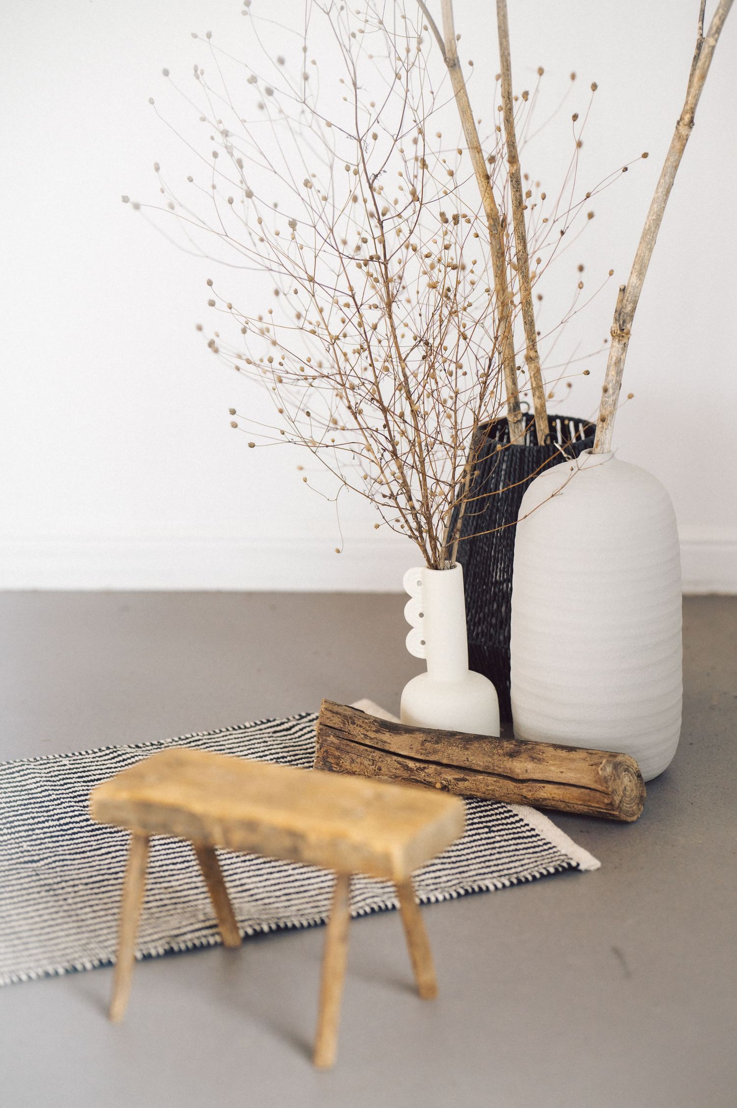 Minimalist decor arrangement with wooden stool, white ceramic vase, black vase, and dried branches on gray rug.