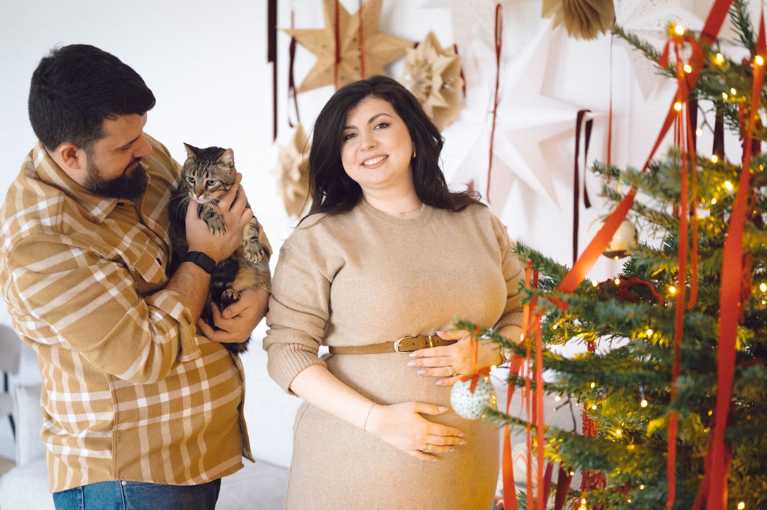 A couple in cozy sweaters decorating a Christmas tree with their cat in a festive living room setting.