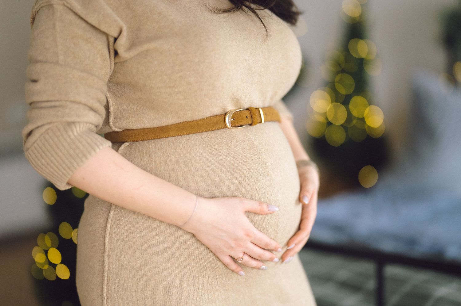A pregnant woman in a beige sweater dress with a brown belt cradles her baby bump with festive lights in the background.