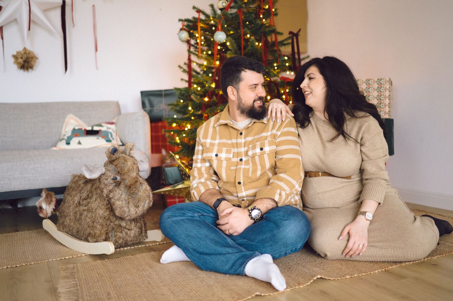 Cat lounging with someone in front of decorated Christmas tree with twinkling lights in cozy living room setting.