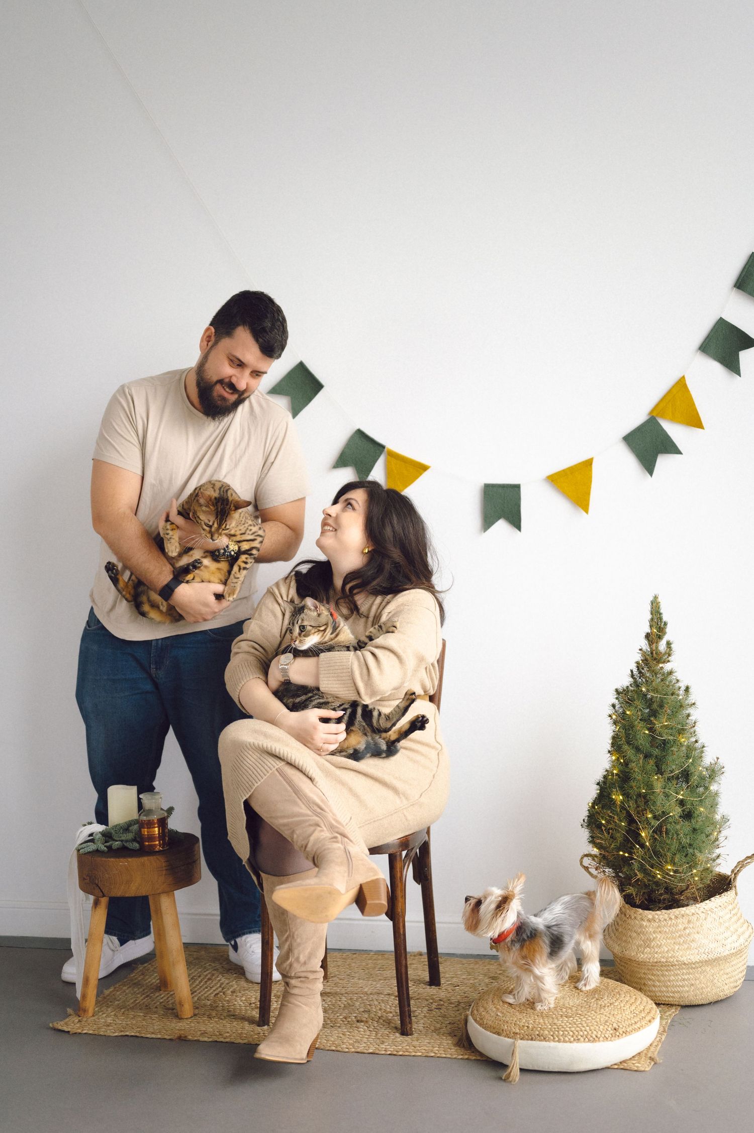 A couple sits with their pets during a holiday photo shoot with festive garlands and a small Christmas tree in the background.