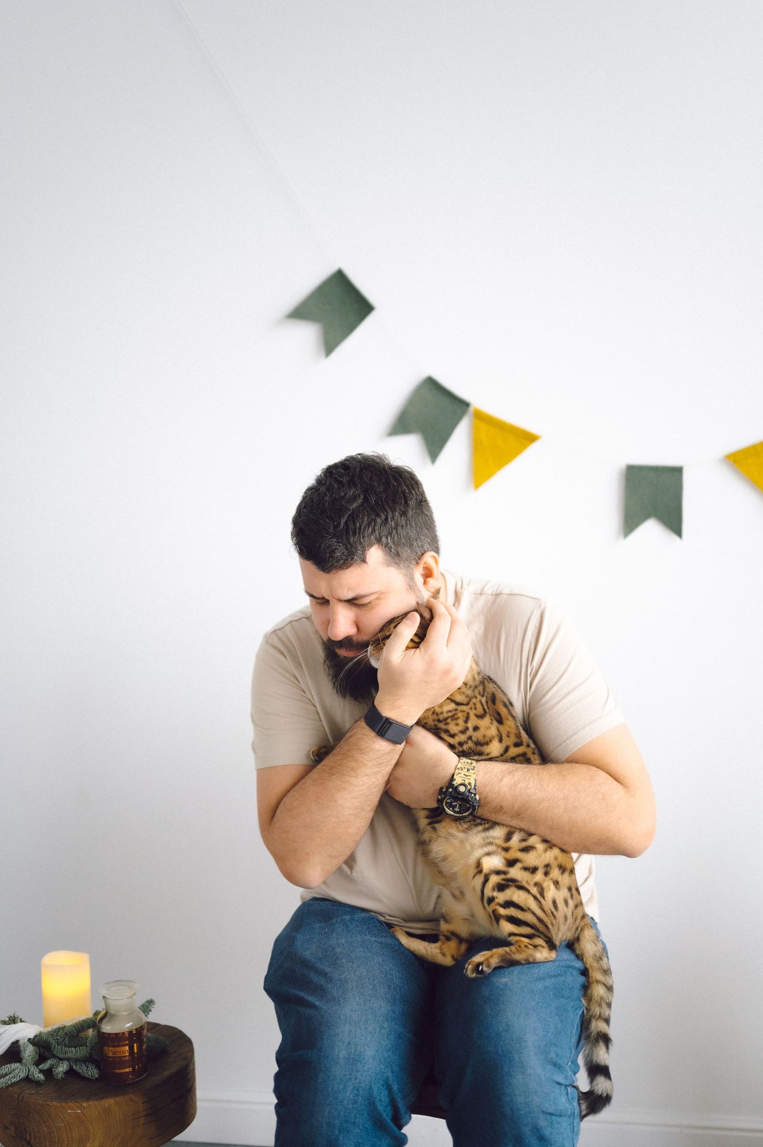 Man sitting on chair holds and comforts a Bengal cat showing signs of distress in a minimalist setting.