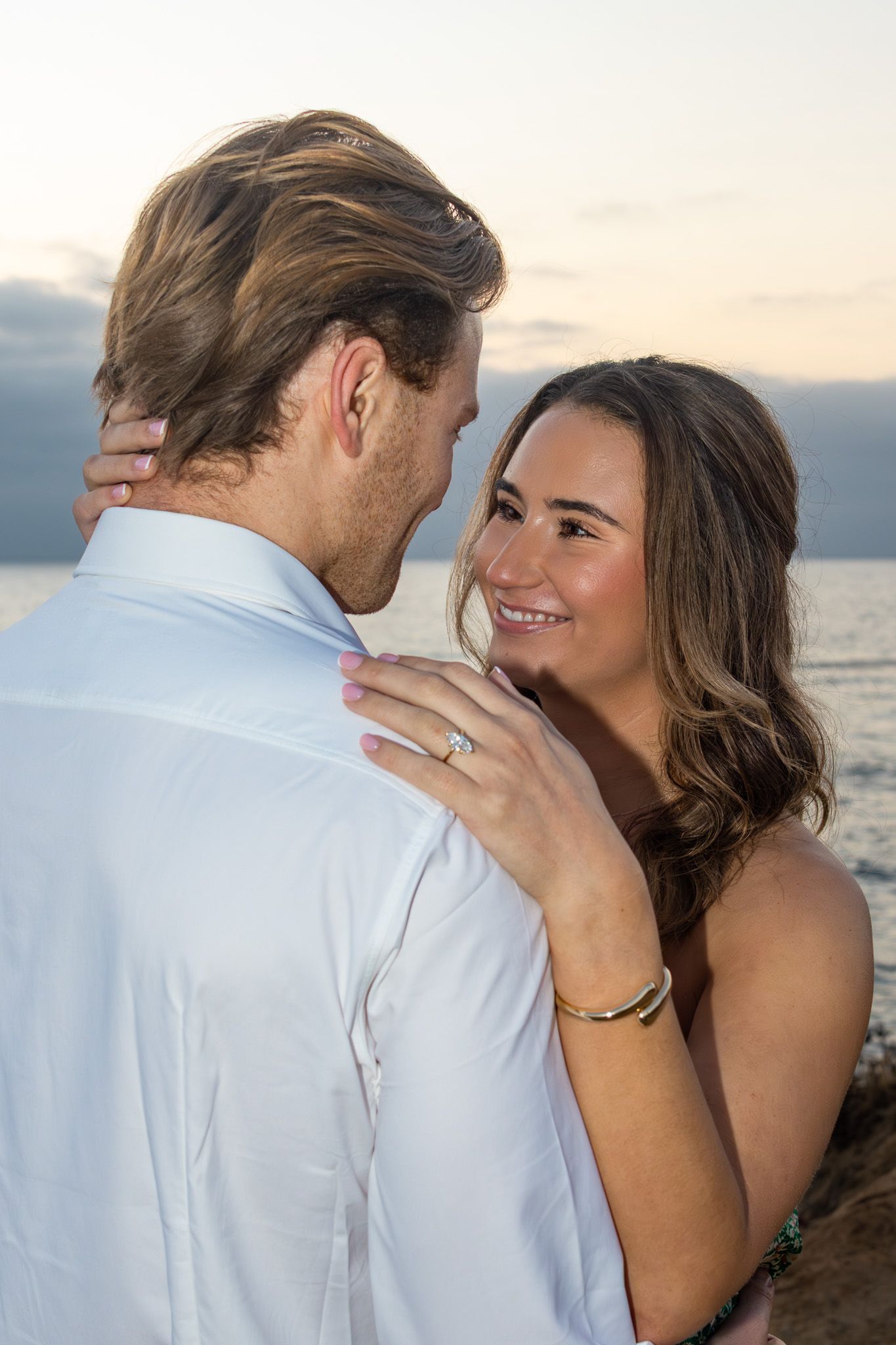 San Diego proposal photography of an engaged couple embracing at Sunset Cliffs, showing off her diamond engagement ring.