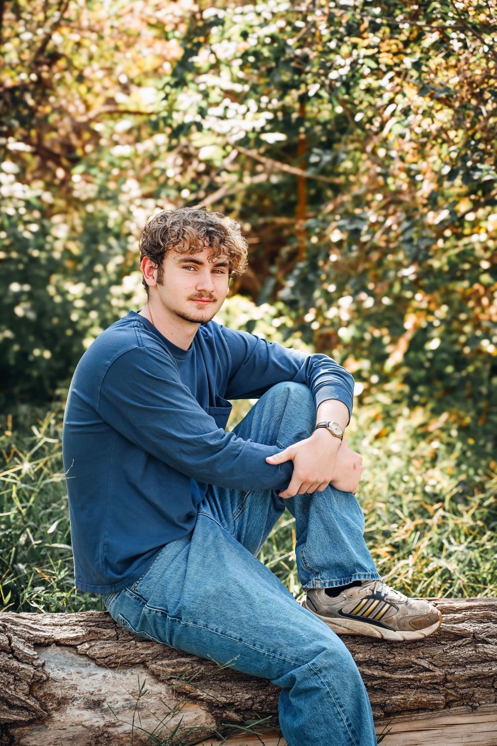 Young man with curly hair sitting on a log in a lush green forest, wearing blue jeans and a navy sweater.