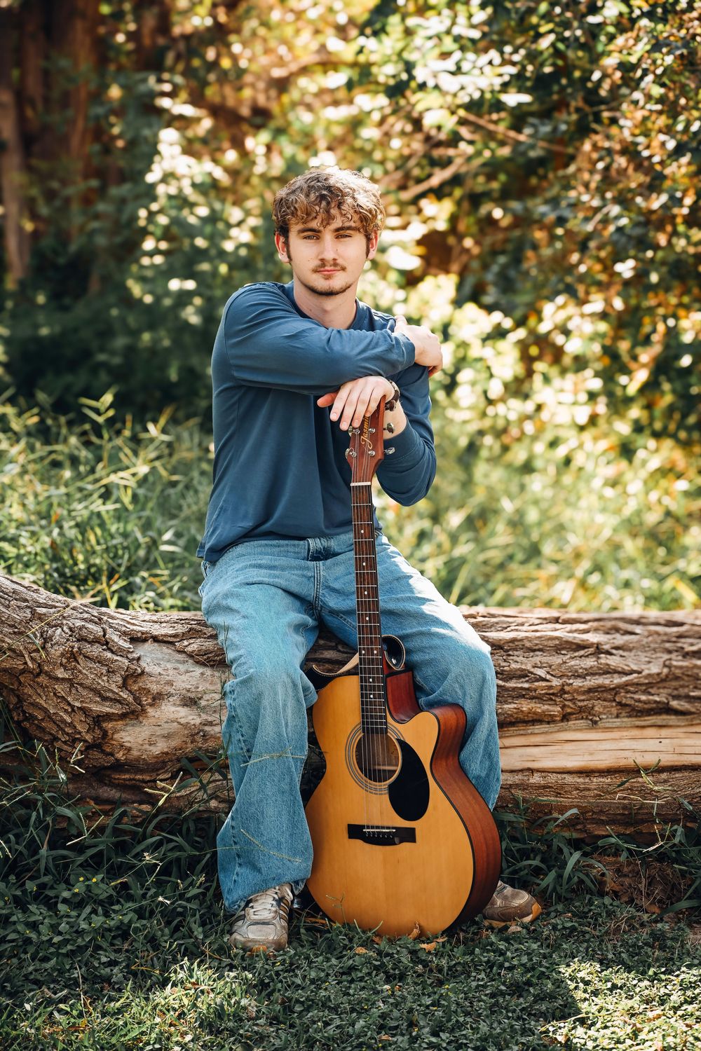Young man with curly hair sits on a log outdoors, leaning on an acoustic guitar in a lush green forest setting.