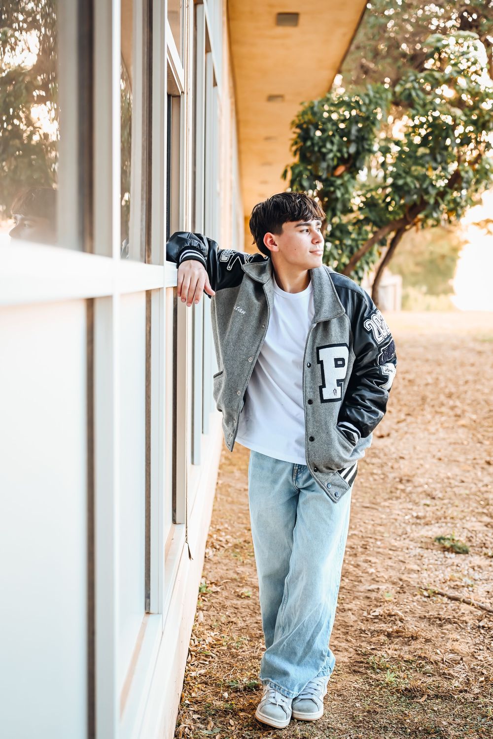 Teen boy in varsity jacket and jeans leans against white building wall outdoors, looking away thoughtfully.