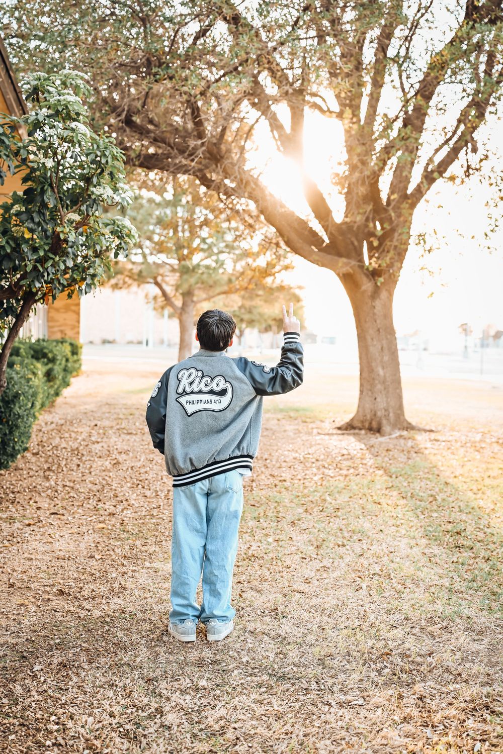 Person in Rico varsity jacket waving under sunlit trees on a leaf-covered path.