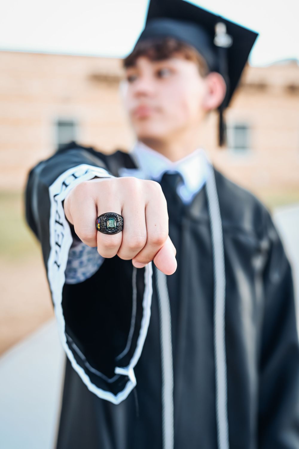 Graduate in black cap and gown proudly displays class ring toward camera in celebratory outdoor photo.