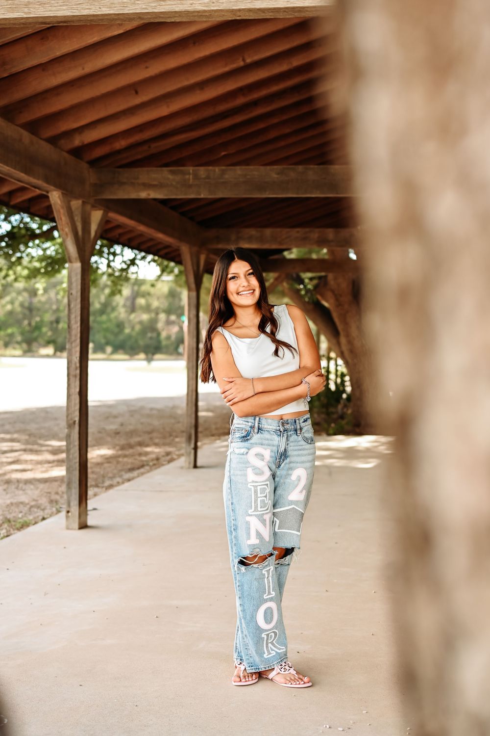 A smiling high school senior poses in ripped jeans with "Senior 25" written on them under a wooden pavilion.