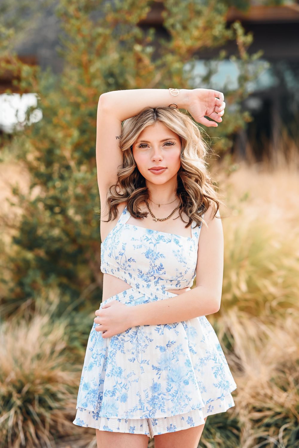 Young woman in blue floral romper poses outdoors among golden grasses and green shrubs in warm natural light.
