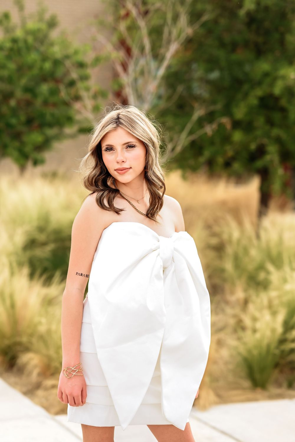Young woman in white bow dress poses outdoors with golden grass and green trees in background.