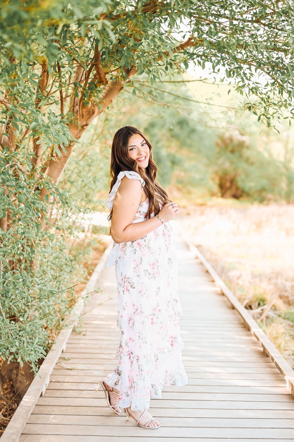 Pregnant woman in floral maxi dress smiling on wooden boardwalk surrounded by lush green trees in soft natural light.