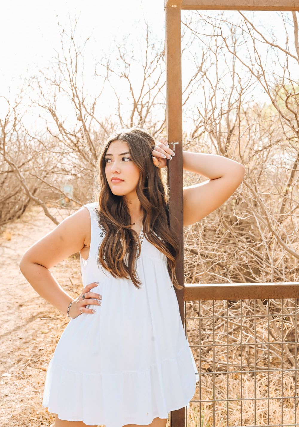 Young woman in white dress poses by rustic metal gate surrounded by bare trees in warm golden light.