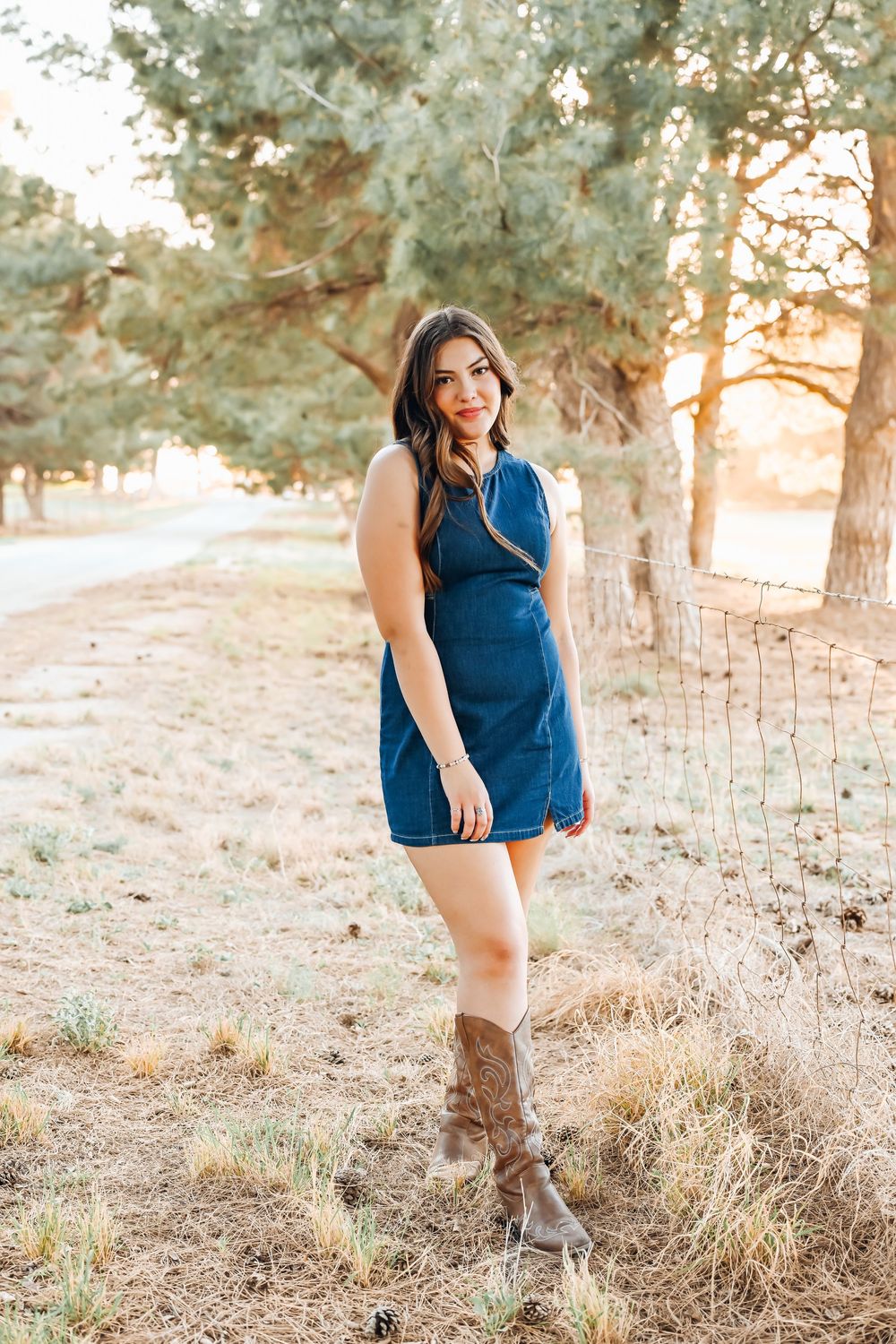 Young woman in denim dress and cowboy boots posing outdoors near pine trees at golden hour.