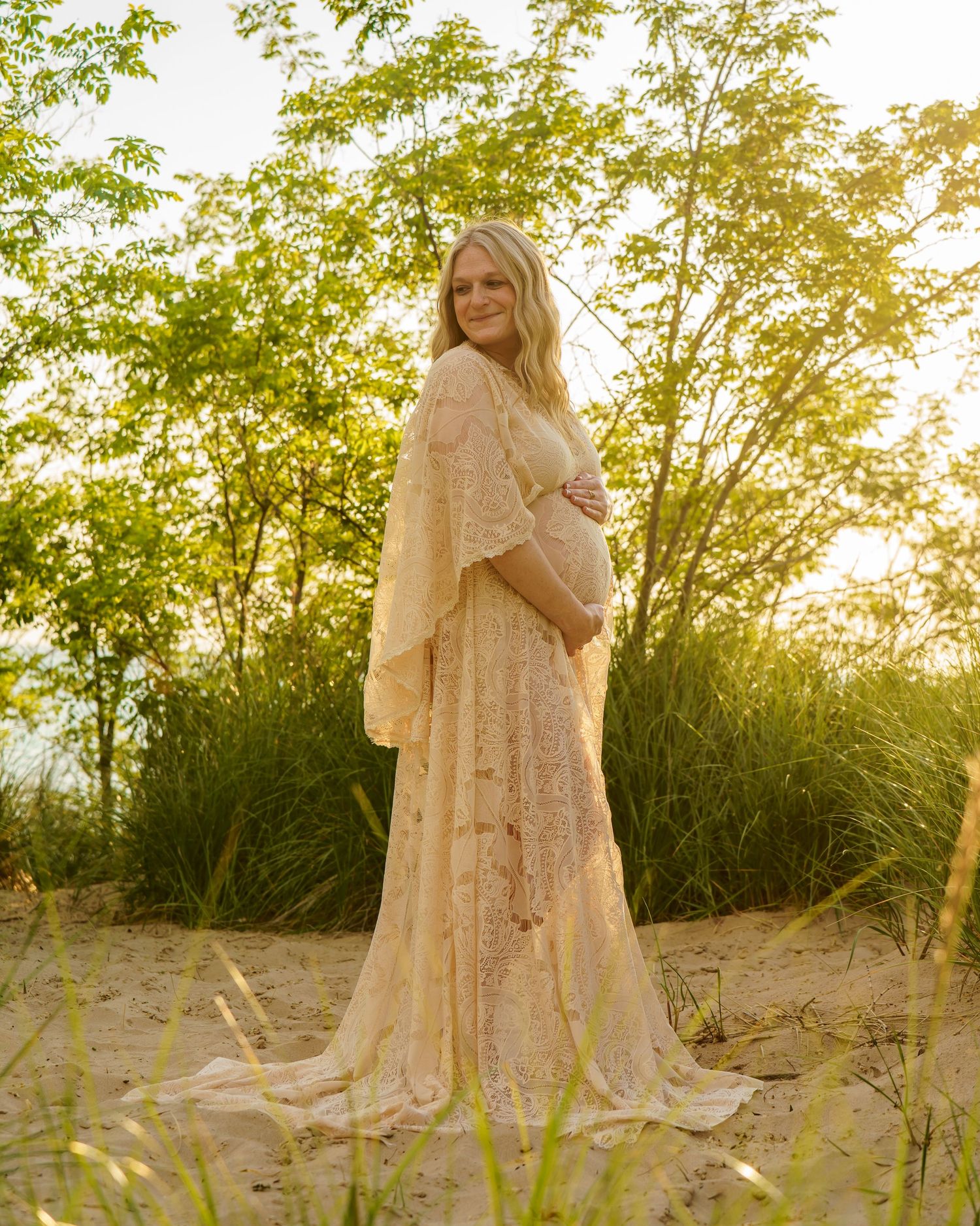 Pregnant woman in elegant lace maternity gown standing on sandy beach surrounded by lush green trees at golden hour.