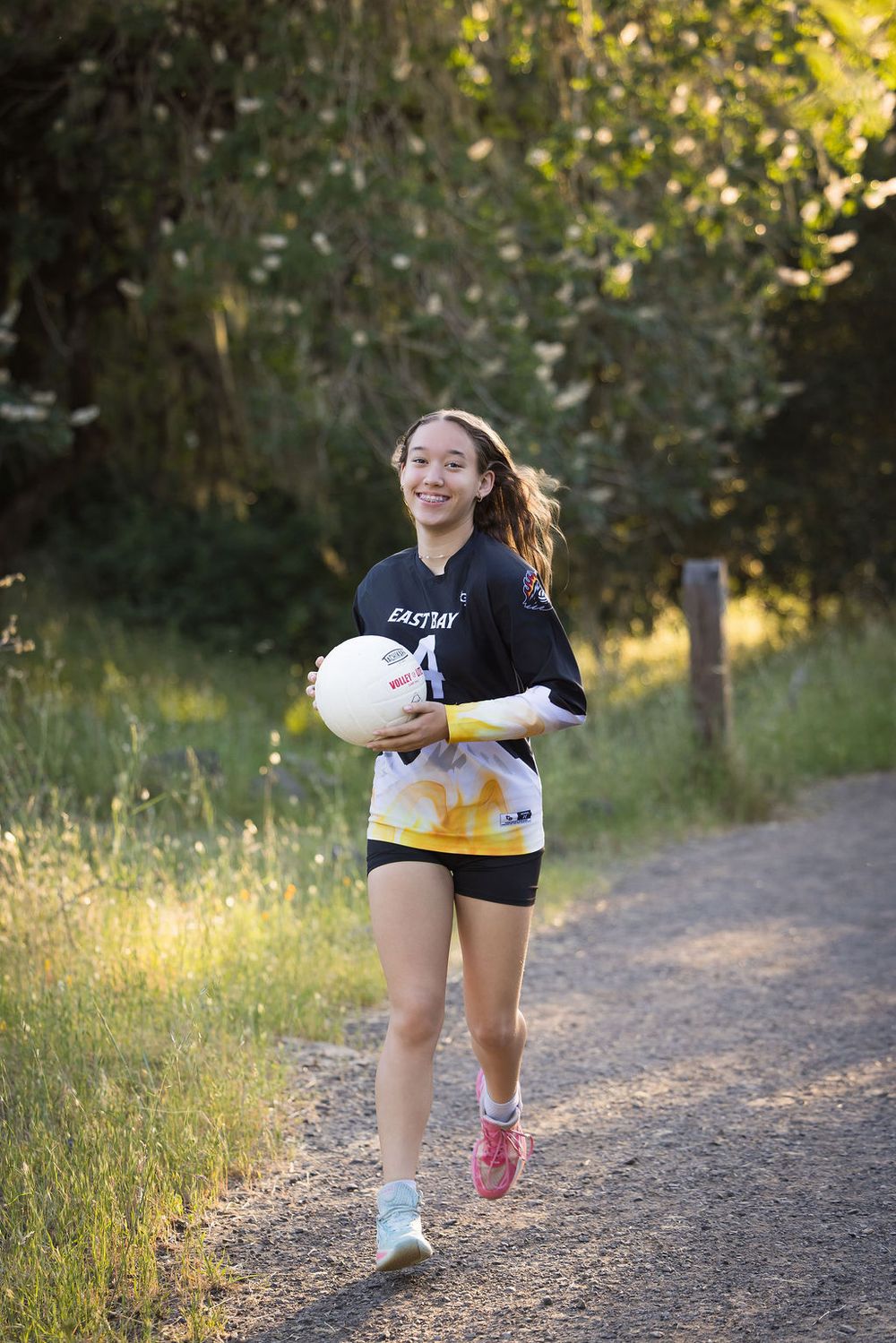 Young girl in black Eastside volleyball jersey smiles while carrying a ball down a sunlit outdoor path.