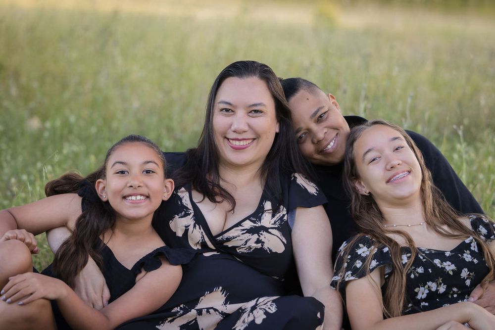 A mother and her three daughters smile together in a sunlit green field, wearing matching black outfits.