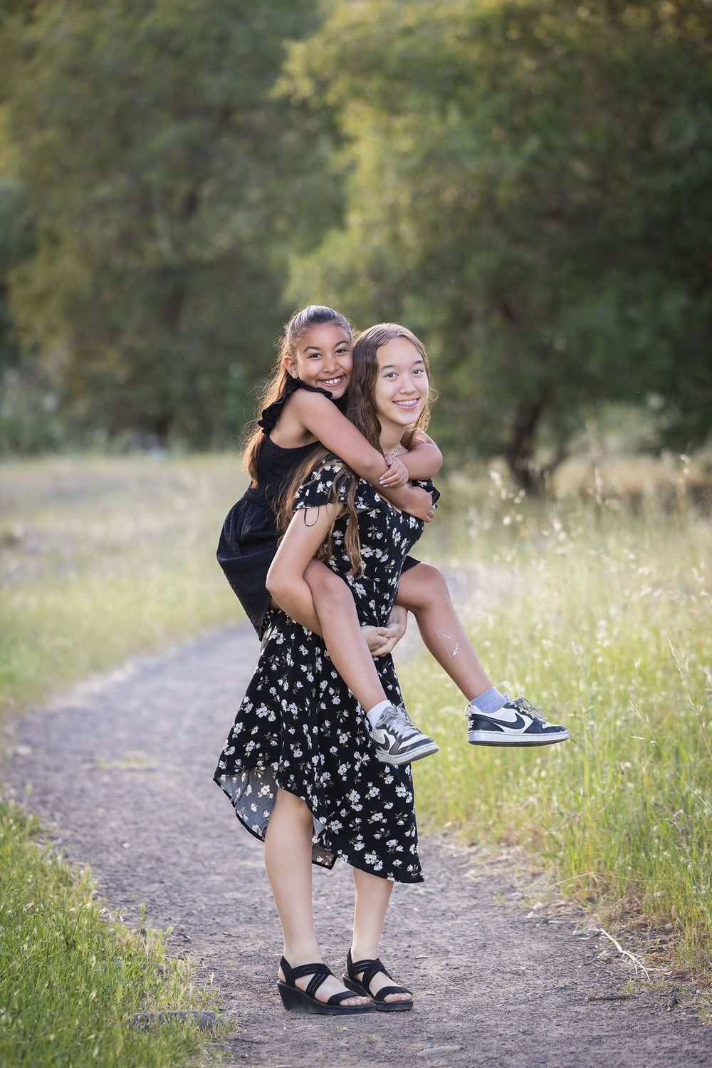 Two girls in black floral dresses laugh while one gives the other a piggyback ride on a scenic outdoor path.