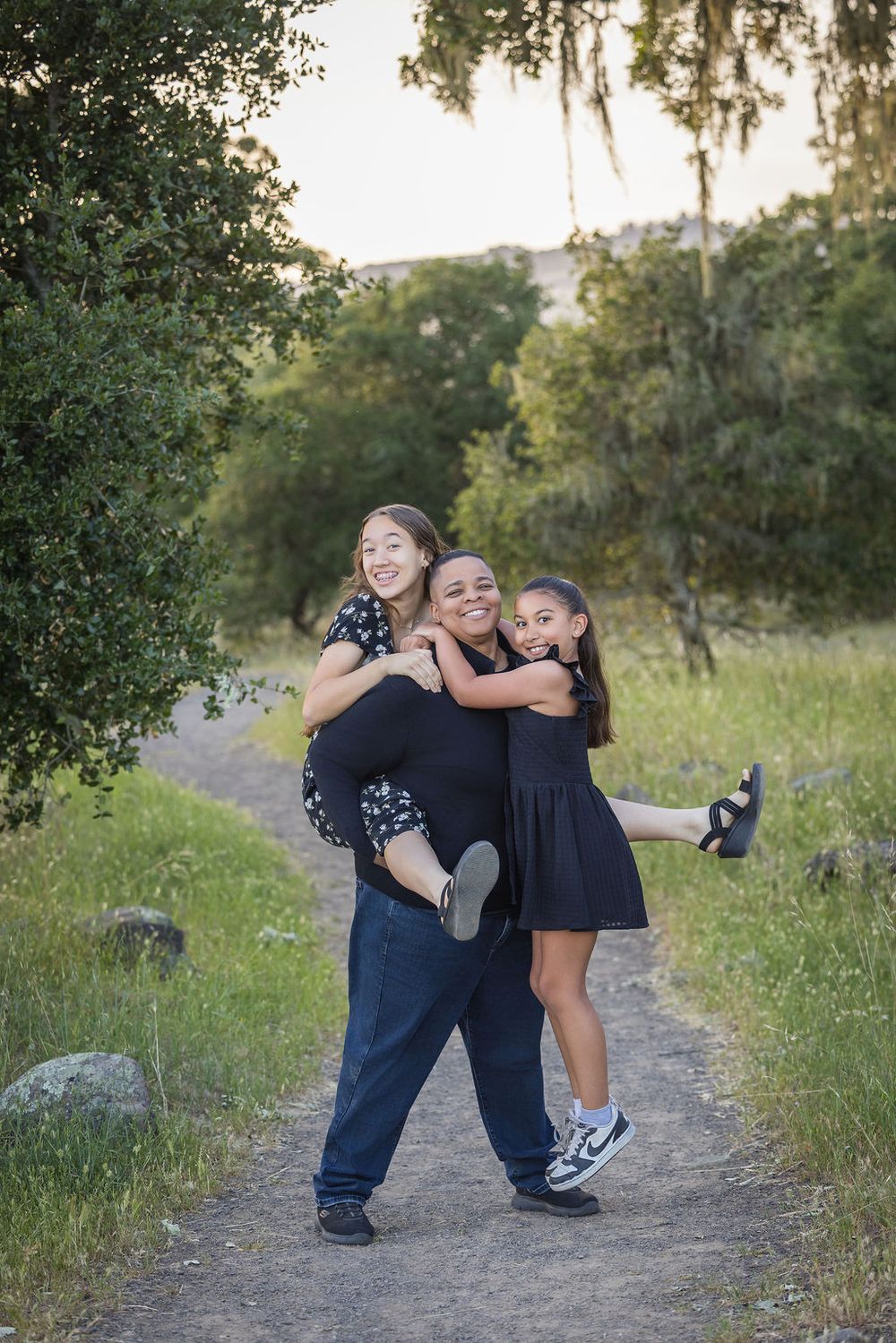 A joyful woman carries two laughing girls on a scenic dirt path surrounded by lush green trees and hills.