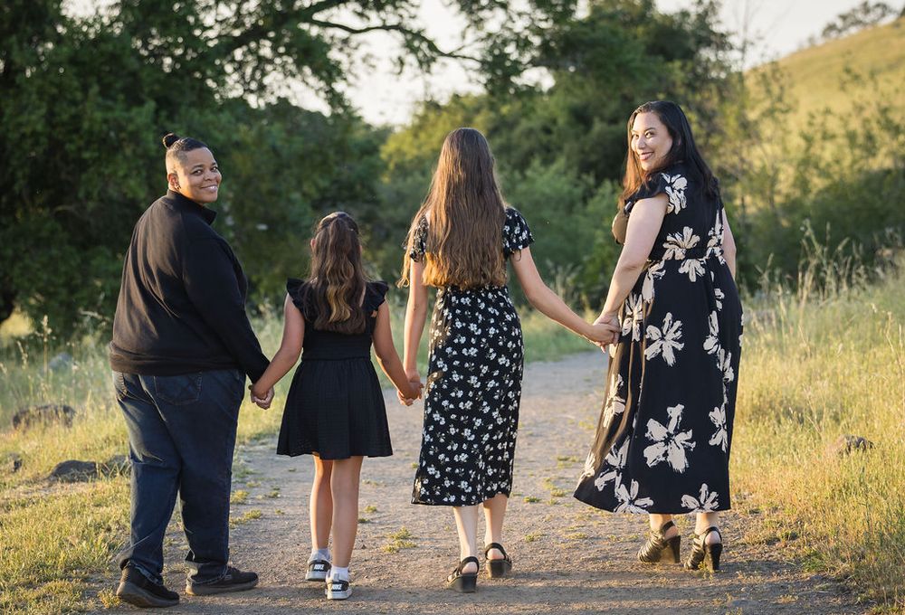 Family of four holding hands walking down a sunlit dirt path surrounded by green trees and golden grass.