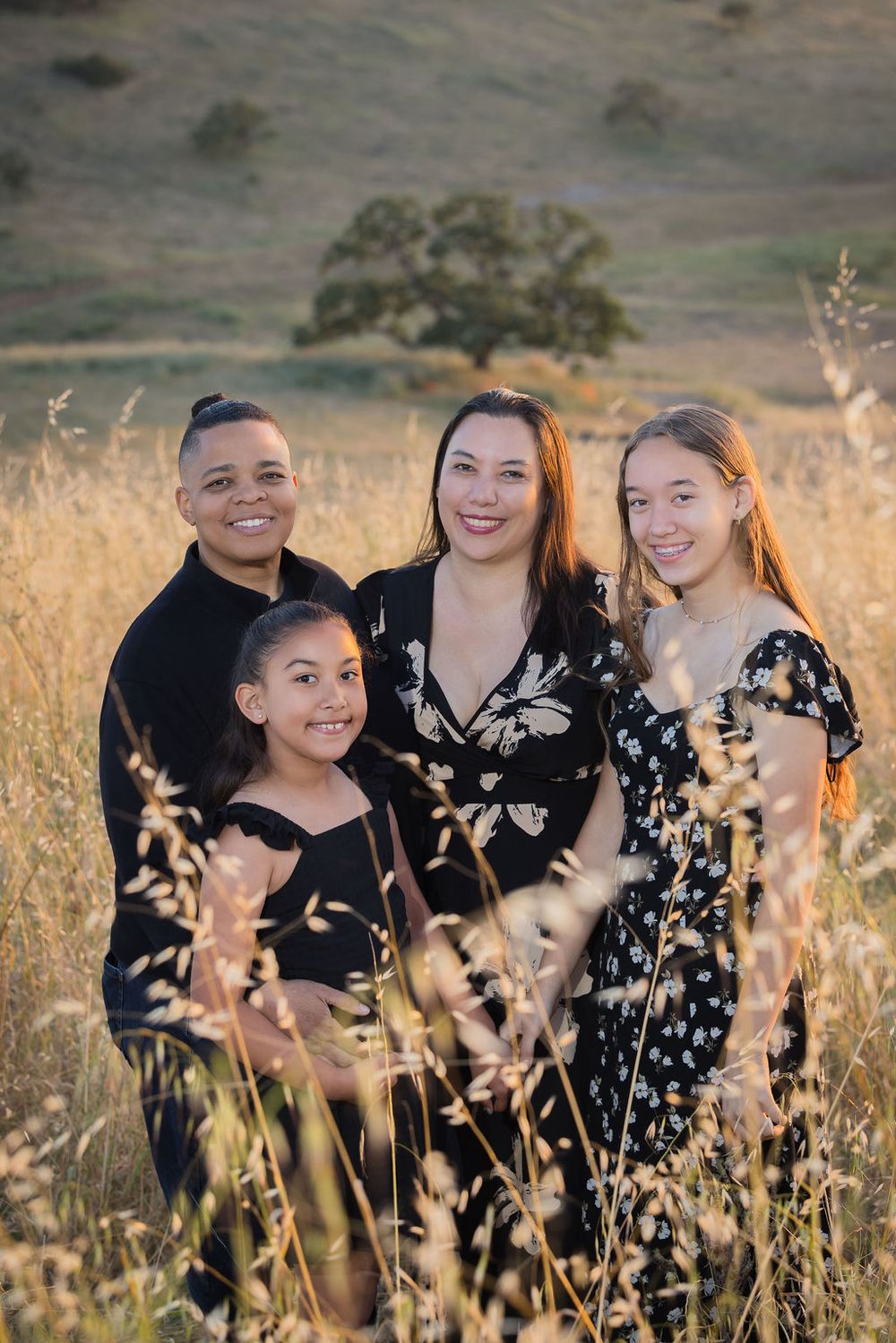 Family of four posing in golden field at sunset, smiling together surrounded by tall grass with rolling hills behind.