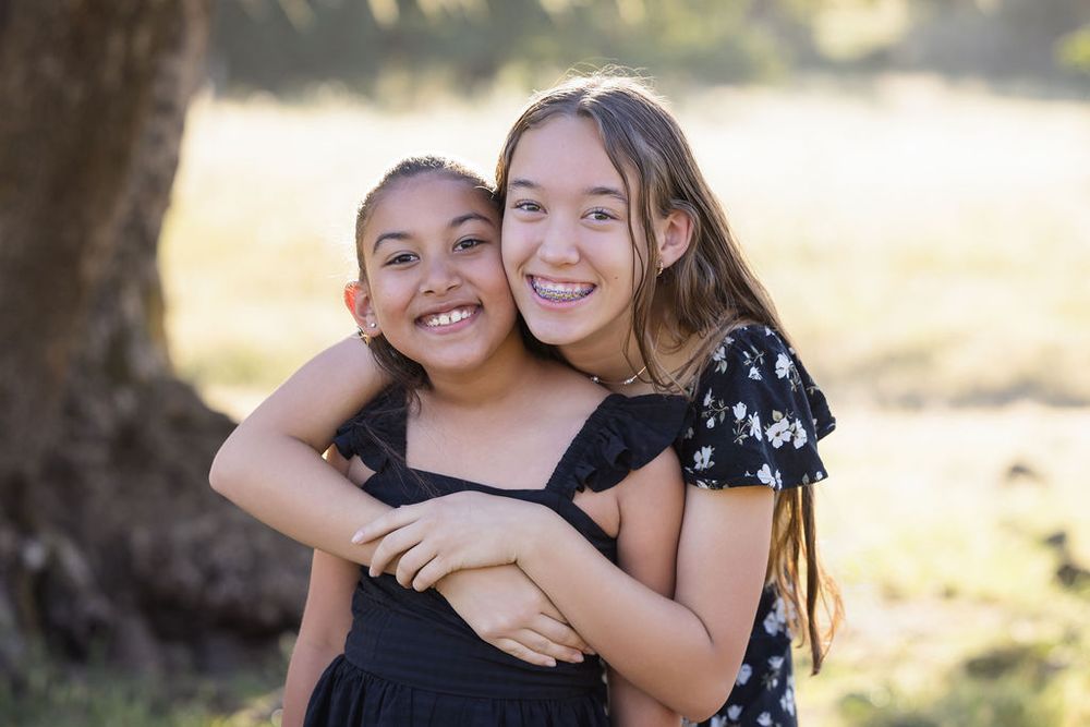 Two smiling girls hugging outdoors in a sunny park, wearing black dresses, sharing a joyful moment together.
