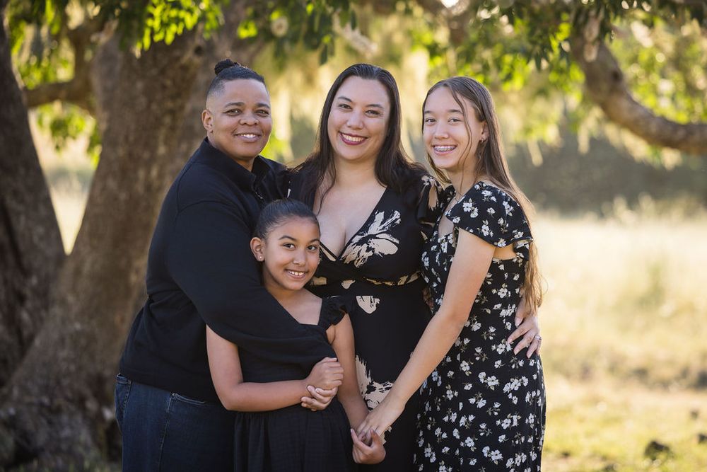 Four women and girls smile together outdoors under a tree in a warm, sunlit family portrait.