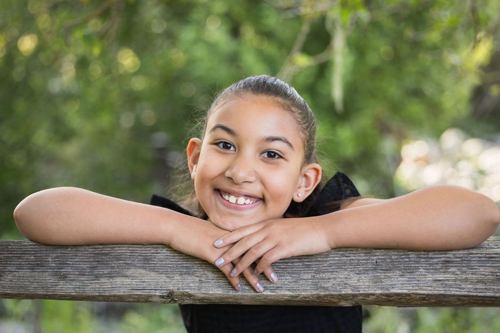 Smiling young girl resting her chin on a wooden fence rail outdoors with green trees in the background.
