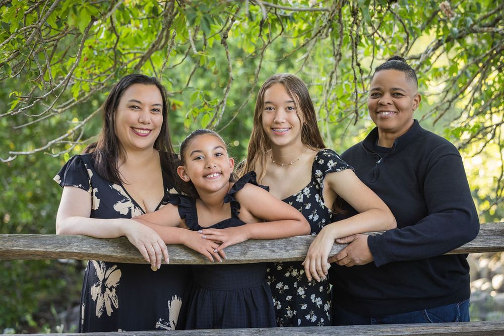 Family of four smiling together on a wooden bridge surrounded by lush green trees in an outdoor park setting.
