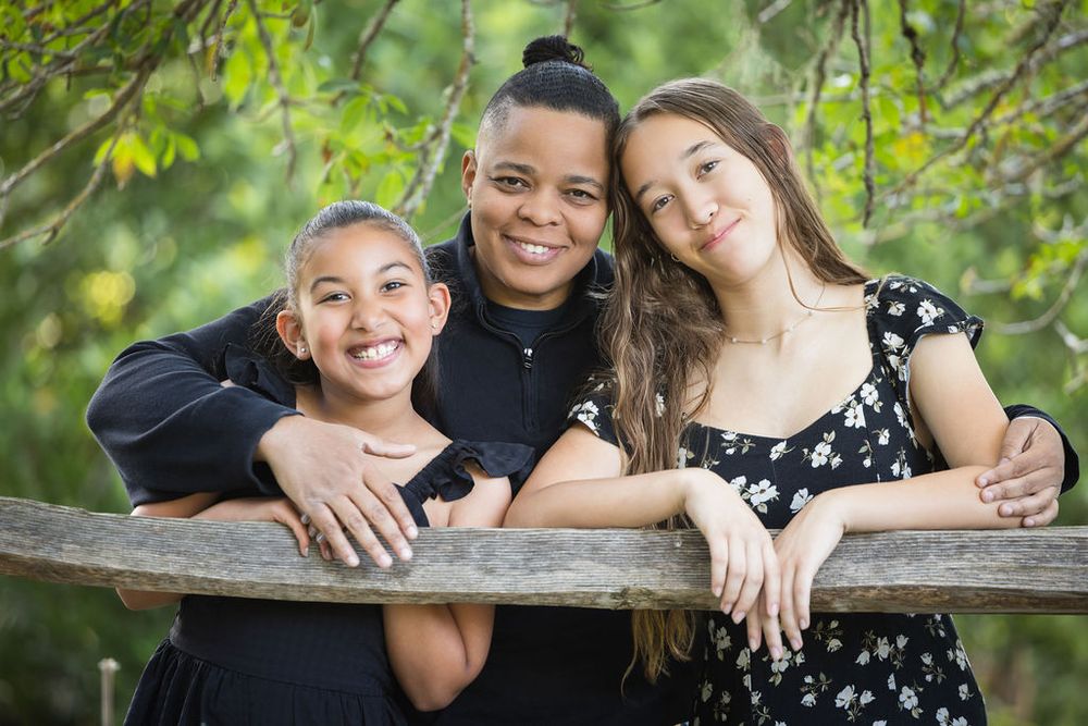 Three smiling children pose outdoors leaning on a wooden fence surrounded by lush green trees.