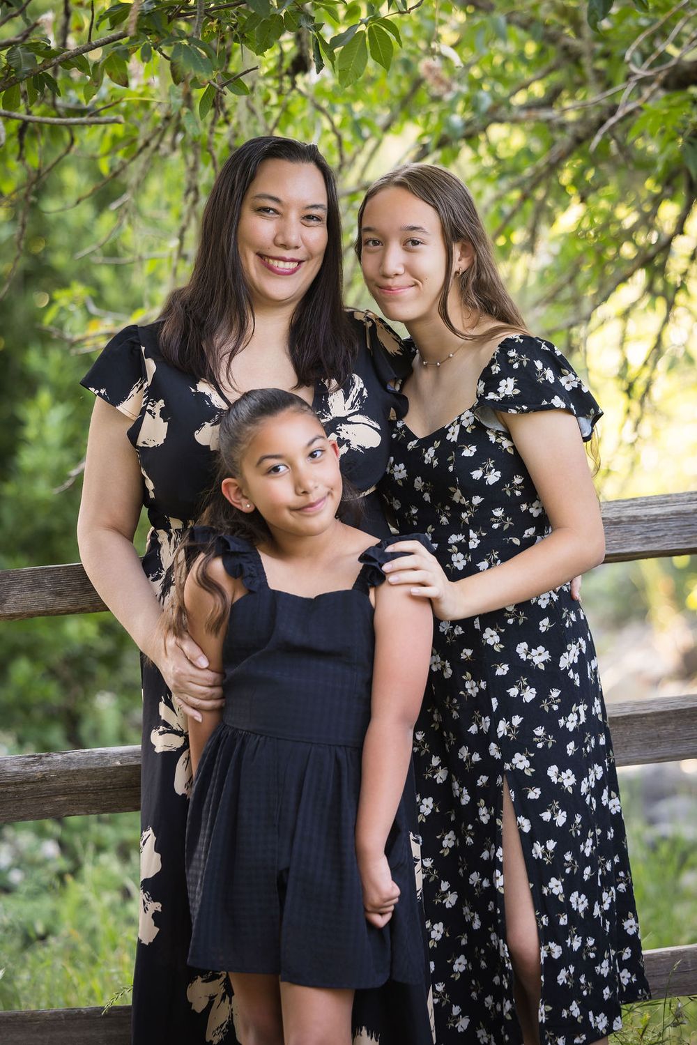 A woman and two girls in matching black floral dresses pose by a wooden fence surrounded by lush green trees.