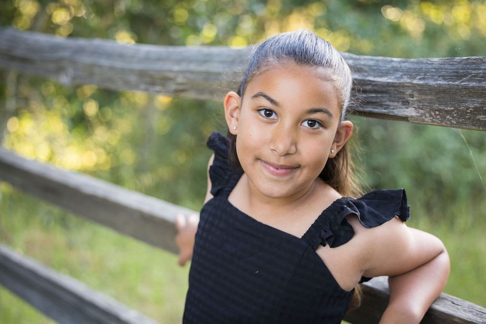 A young girl in a black ruffled top poses by a wooden fence outdoors, smiling softly at the camera.