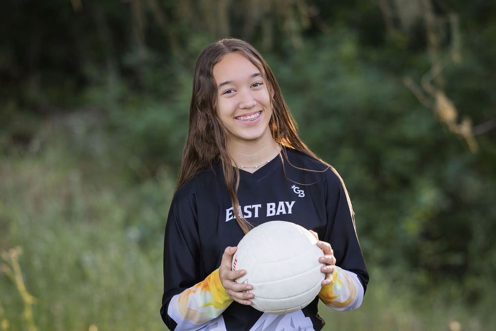 Young girl in East Bay jersey smiling and holding a volleyball outdoors.