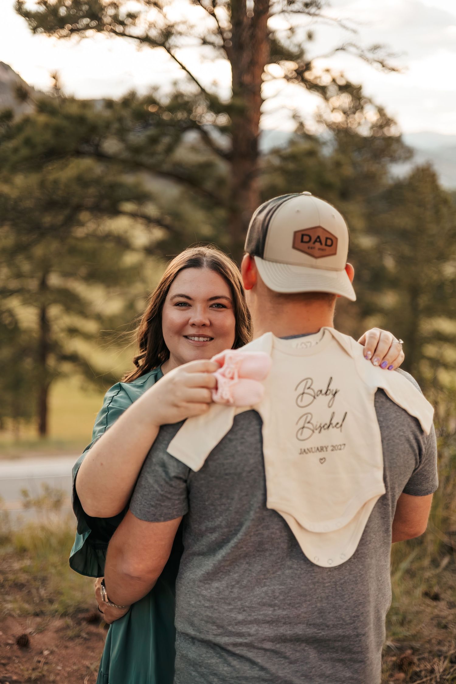 Couple holding a  onesie outdoors, man wearing a Dad hat, woman smiling, p | Baby Announcement Photographer in Boulder Colorado