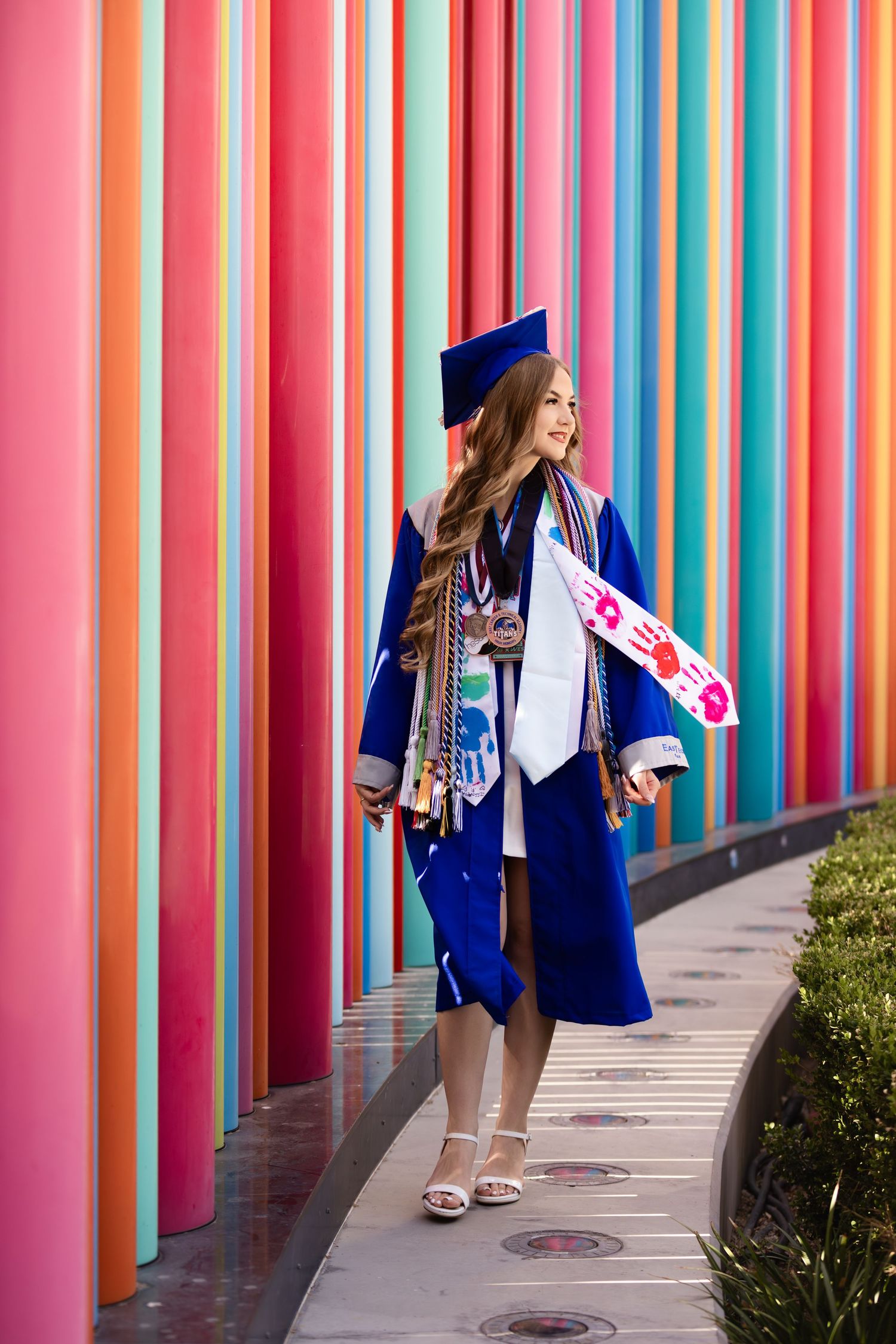 Graduate in blue cap and gown holding diploma in front of colorful striped wall outdoors.