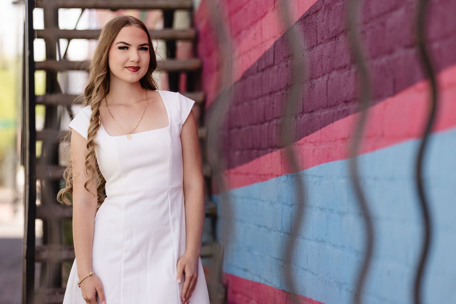Young woman in white dress poses by colorful graffiti wall with pink and blue art in urban setting.