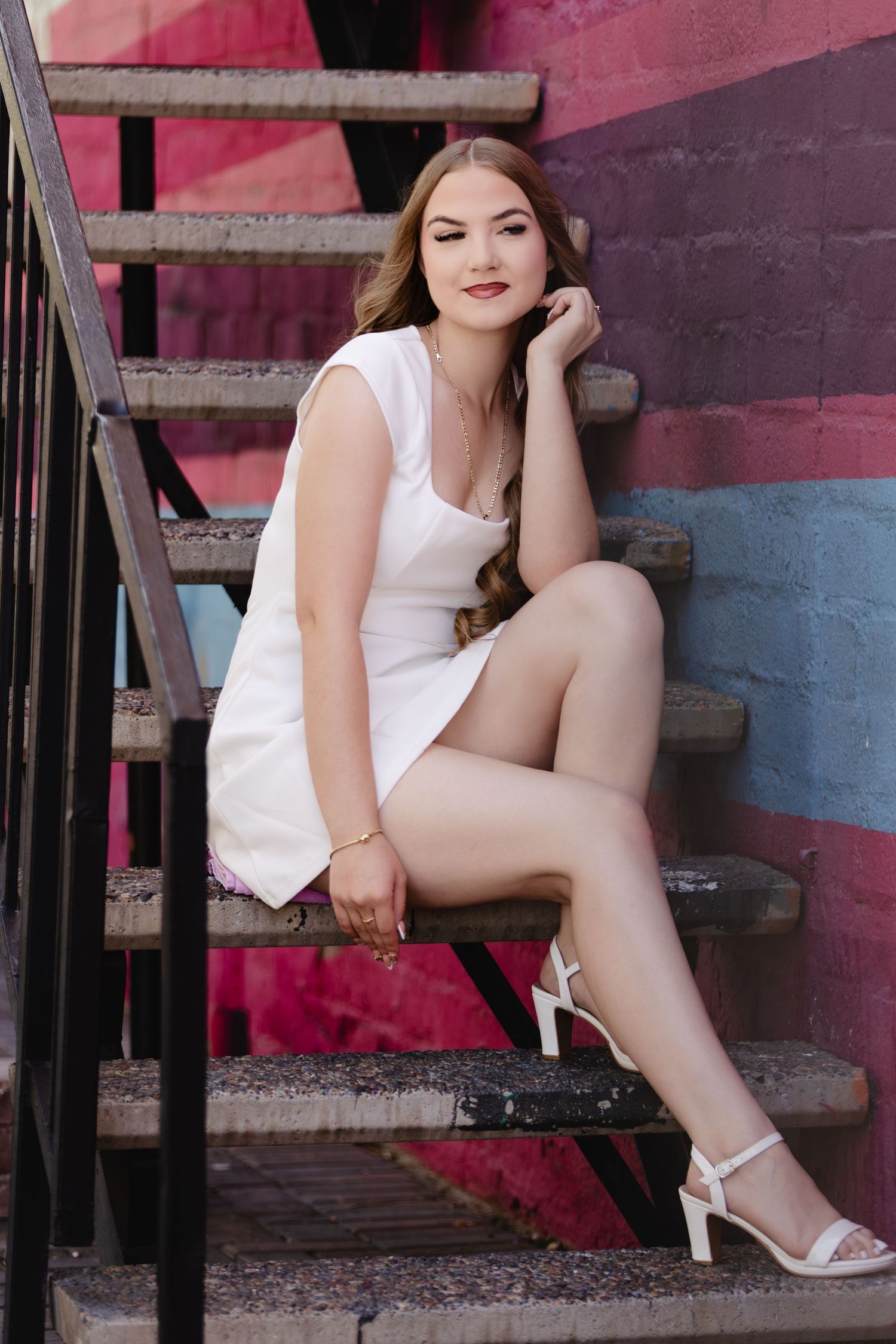 Young woman in white mini dress and heels sitting gracefully on outdoor stairs with colorful mural wall backdrop.