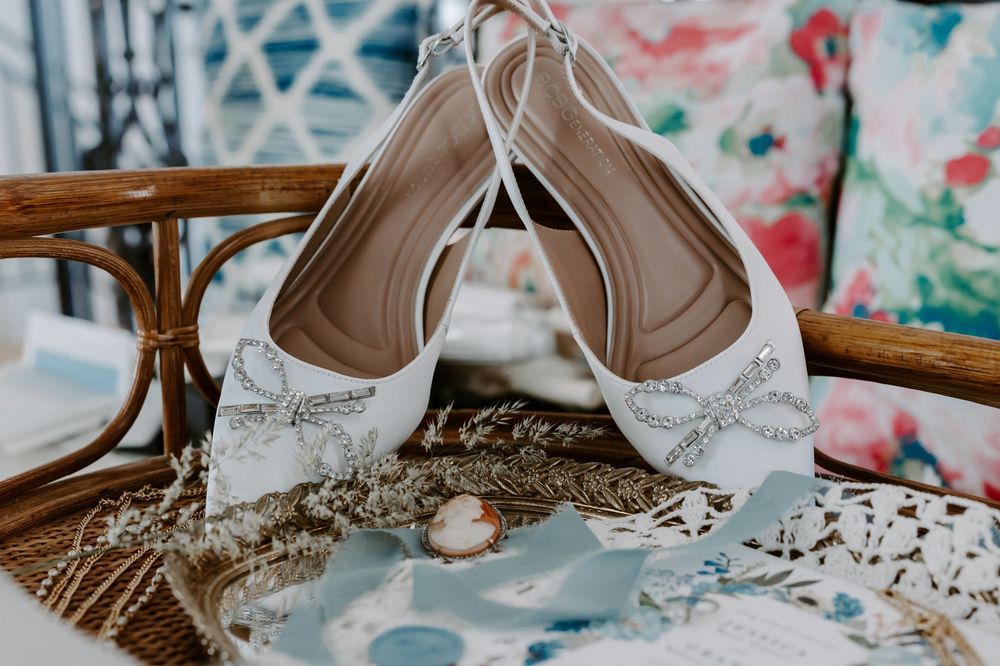 White bridal slingback heels with crystal bow embellishments displayed on a wicker chair.