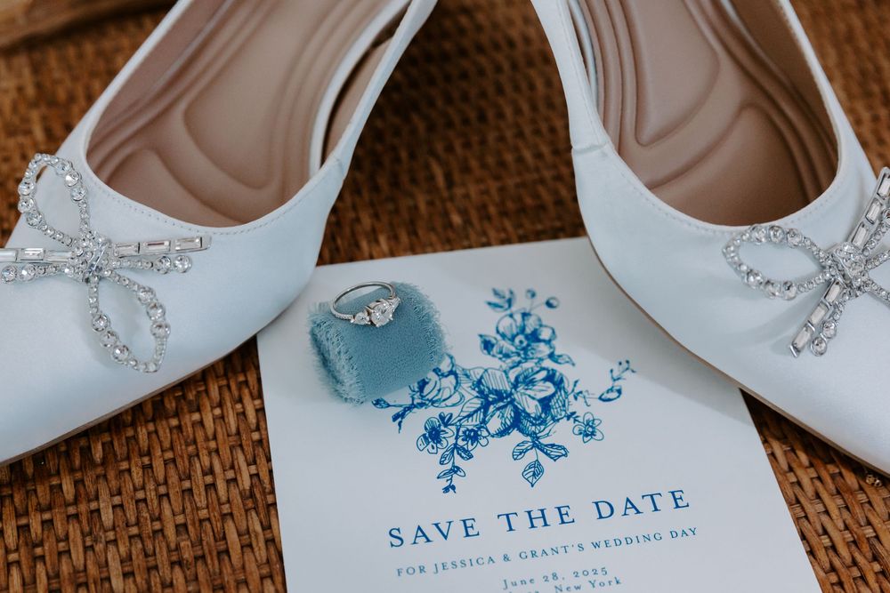 White bridal heels with crystal bows resting on a blue floral Save the Date card with a ring box.